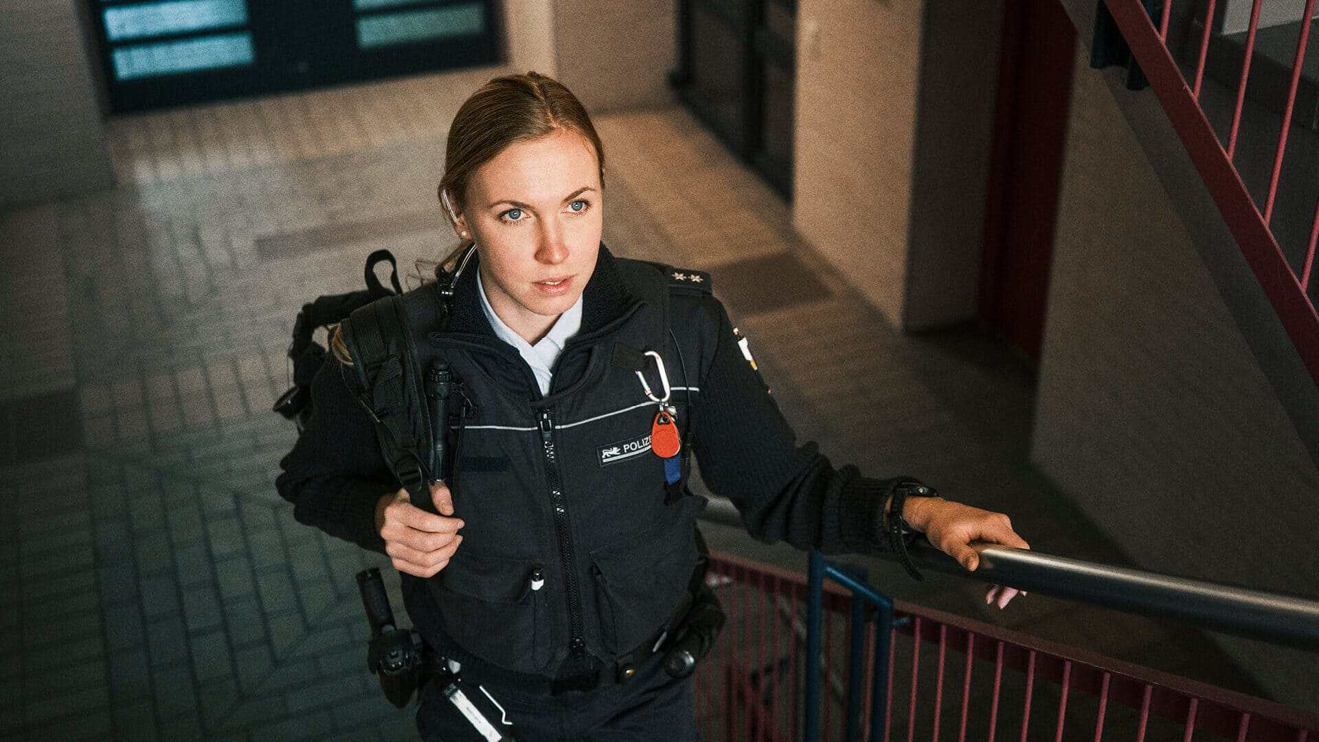 Mitarbeiterin Vivien, die mit einem Rucksack auf der Schulter in eine Polizeistation die Treppen hoch läuft. Im Hintergrund sind Türen zu sehen. Sie trägt eine typische Polizeiuniform.