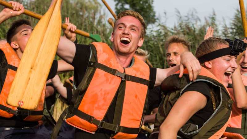 Fünf Jugendliche, drei Jungen und zwei Mädchen, sind auf einem Fluss oder einem See zu sehen. Sie tragen alle leuchtend orangefarbene Schwimmwesten und haben Spaß bei einer gemeinsamen Aktivität.