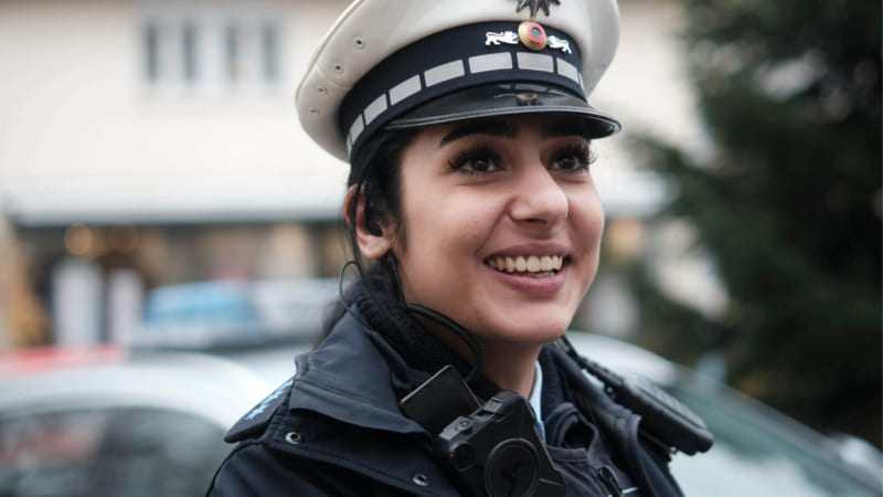 Eine Polizistin in Uniform lächelt in die Kamera. Im Hintergrund ist verschwommen ein Polizeiauto zu sehen, möglicherweise das Fahrzeug, mit dem die Polizistin unterwegs ist. Hinter dem Auto befindet sich ein Gebäude, das nicht genau zu erkennen ist. Die Polizistin hat eine freundliche Ausstrahlung und wirkt zugänglich und vertrauenswürdig. Das Bild suggeriert, dass die Polizei freundliches und hilfsbereit ist, das stets für die Sicherheit der Bevölkerung sorgt.